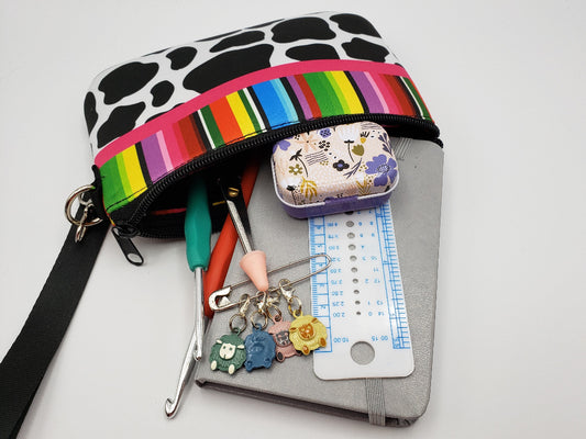 Cow print pouch with rainbow strap on a gray surface, containing sewing tools and accessories.