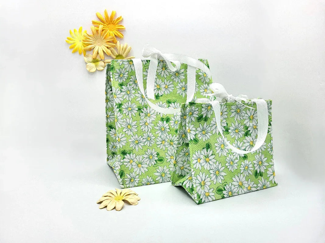 Two green tote bags with white floral pattern and white handles, surrounded by decorative yellow flowers on a white background.