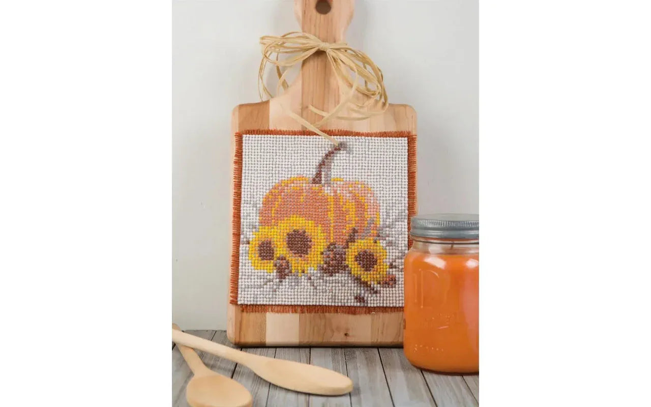 Wooden cutting board with cross-stitched pumpkin design on a wooden surface with a jar of pumpkin puree.